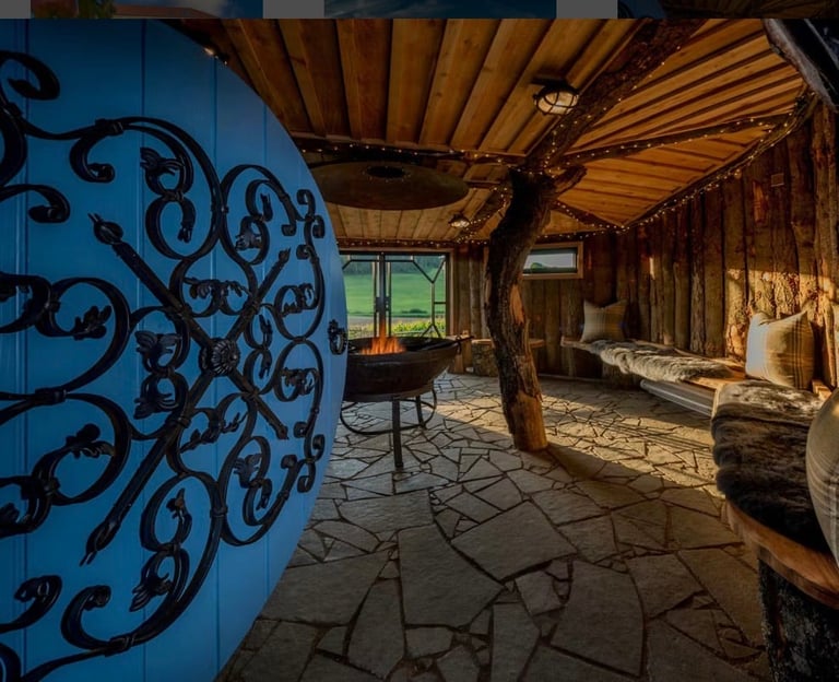 Interior of rustic wooden pavilion with ornate blue door, stone flooring, wooden beams, benches, and window framing countryside view