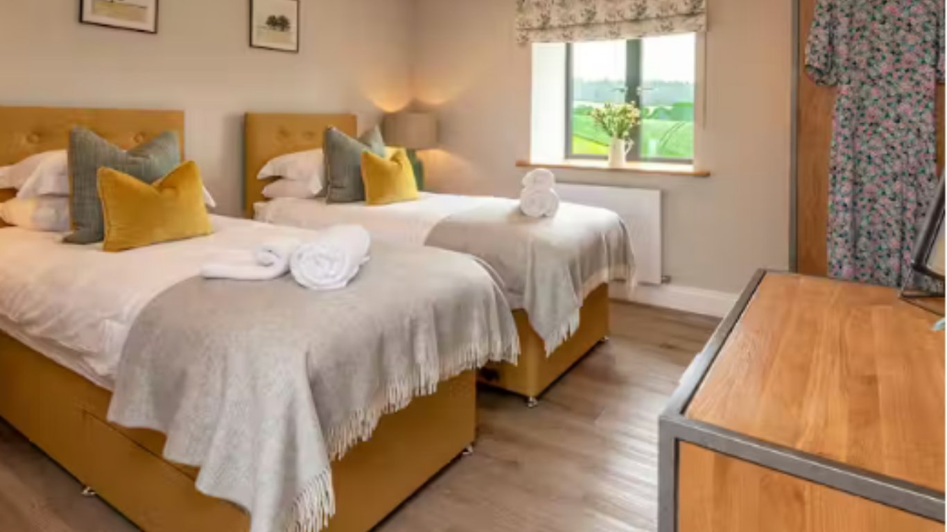 Spacious hotel bedroom with two twin beds in mustard yellow frames, white bedding, yellow pillows, wooden desk, and countryside window view