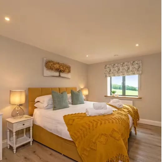 Modern bedroom with gold velvet bed, white bedding, yellow throw blanket, nightstand with lamp, and floral window treatment