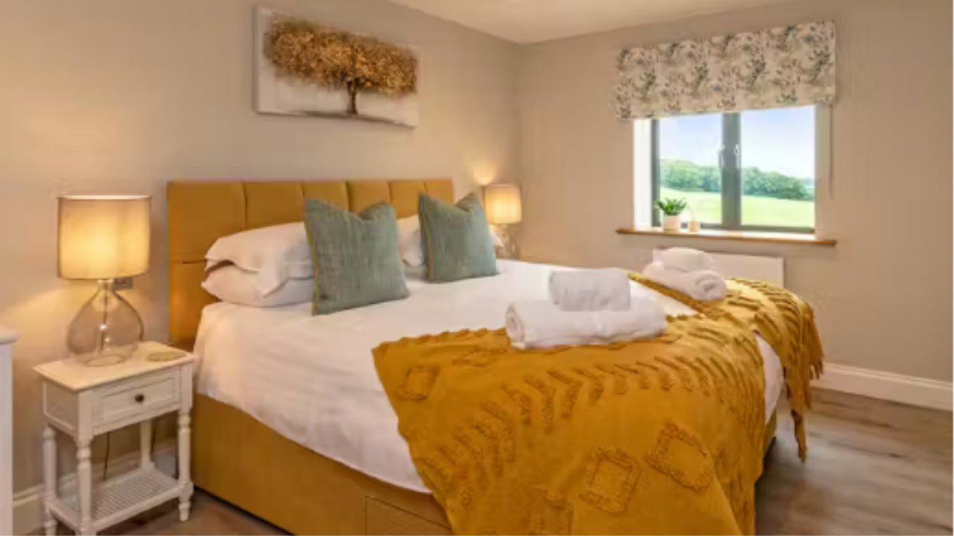 Bright modern bedroom with golden upholstered bed, white linens, green and cream pillows, mustard throw blanket, and white nightstand with lamp