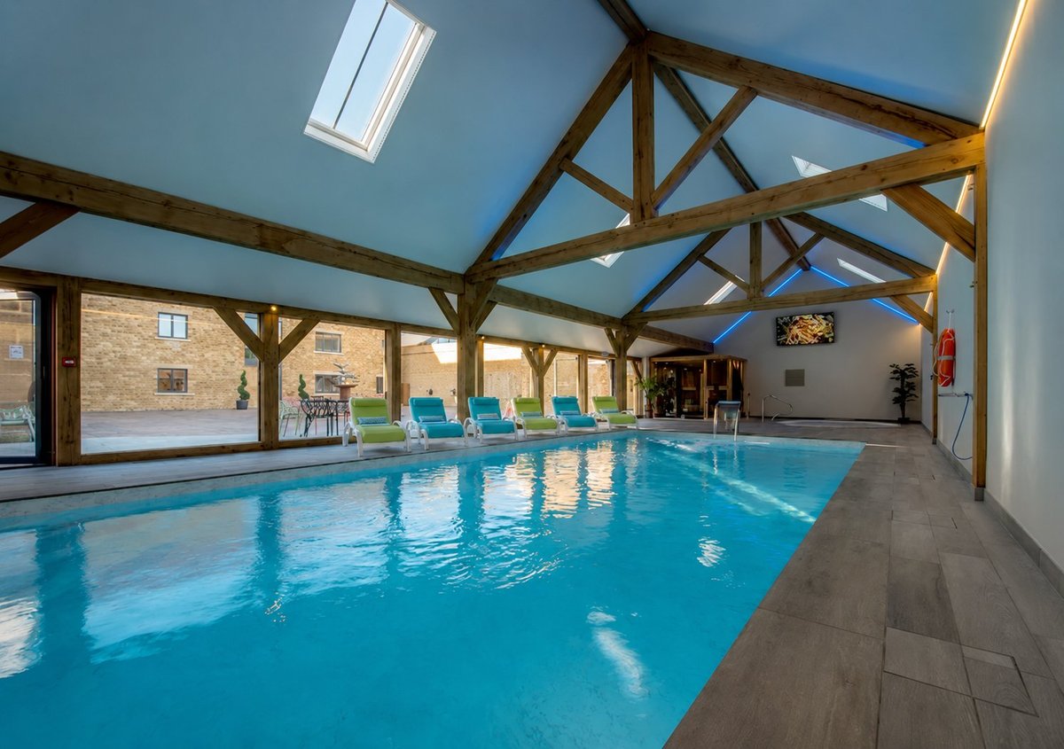 Indoor swimming pool with bright blue water, exposed wooden beam ceiling, large windows, and slate flooring in a modern facility.