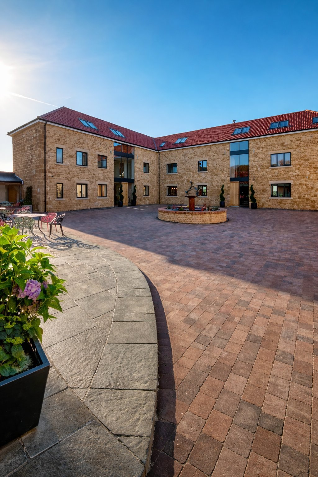 Modern brick courtyard building with red roof, burgundy paved courtyard, planted flowers in foreground, clear blue sky