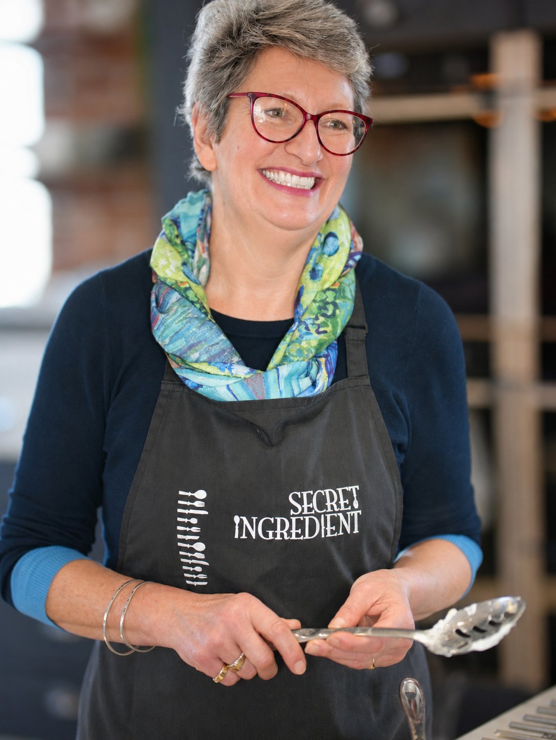 Woman with gray hair and glasses wearing a Secret Ingredient apron, smiling while holding a cooking utensil