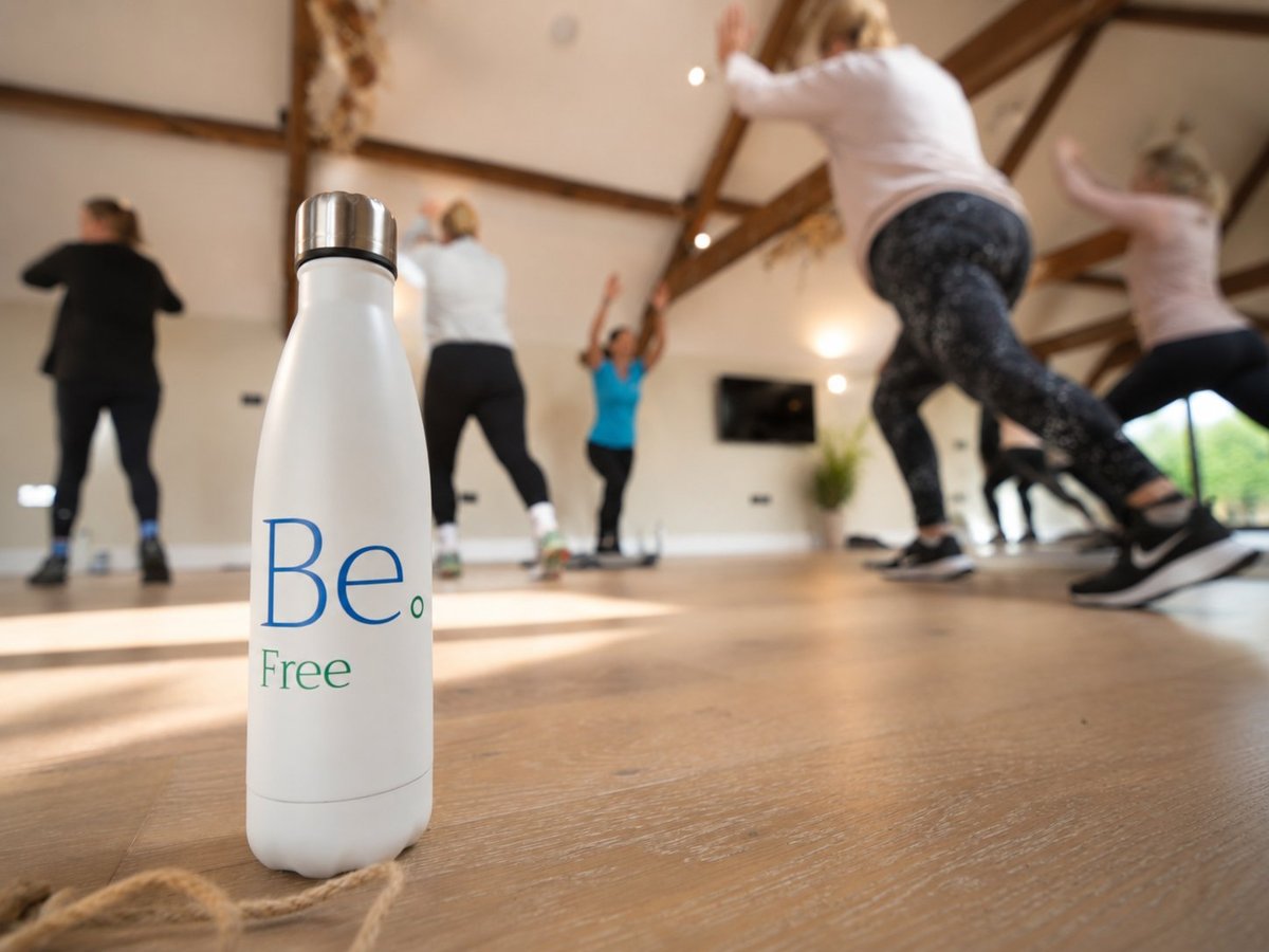White Be Free water bottle on wooden floor with fitness class participants exercising in background